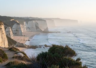 À l’aube, un rivage français calme avec des falaises de calcaire, une plage et un sentier côtier sinueux baignés de lumière douce.