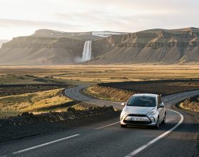 Route islandaise traversant une vaste plaine volcanique, avec une voiture de location discrète, des montagnes et une cascade au loin sous la lumière dorée du soleil d'été.