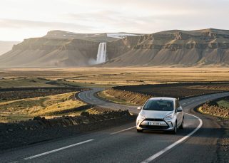 Route islandaise traversant une vaste plaine volcanique, avec une voiture de location discrète, des montagnes et une cascade au loin sous la lumière dorée du soleil d'été.