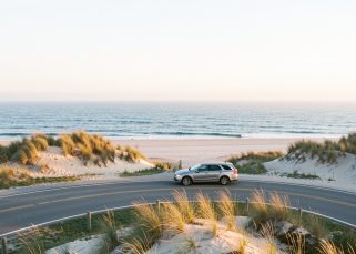 Une voiture discrète roule sur une route côtière de Cape Cod au lever du soleil, entre les dunes blondes et l’océan Atlantique.