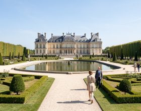 Vue panoramique réaliste du château de Chantilly depuis les jardins à la française, baigné par la lumière claire du matin, avec quelques visiteurs discrets.