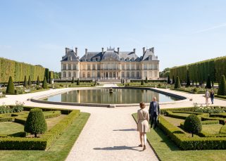Vue panoramique réaliste du château de Chantilly depuis les jardins à la française, baigné par la lumière claire du matin, avec quelques visiteurs discrets.