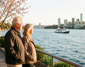 Couple de voyageurs de plus de 50 ans contemplant le port de Sydney au printemps, sous une lumière douce, avec un ferry au loin.